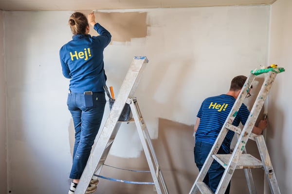 Twee personen schilderen een muur in een lege kamer, elk op een ladder voor een strakke afwerking.