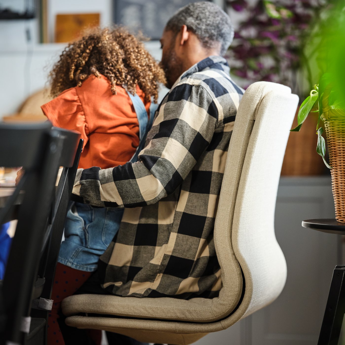 A dining area where a man sits on a beige-white MULLFJÄLLET conference chair next to a dining table, his daughter in his lap.