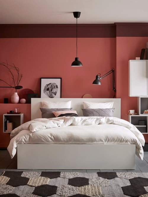 White MALM bed frame with white quilt cover in front of a red bedroom wall next to a light grey wall-mounted shelf.
