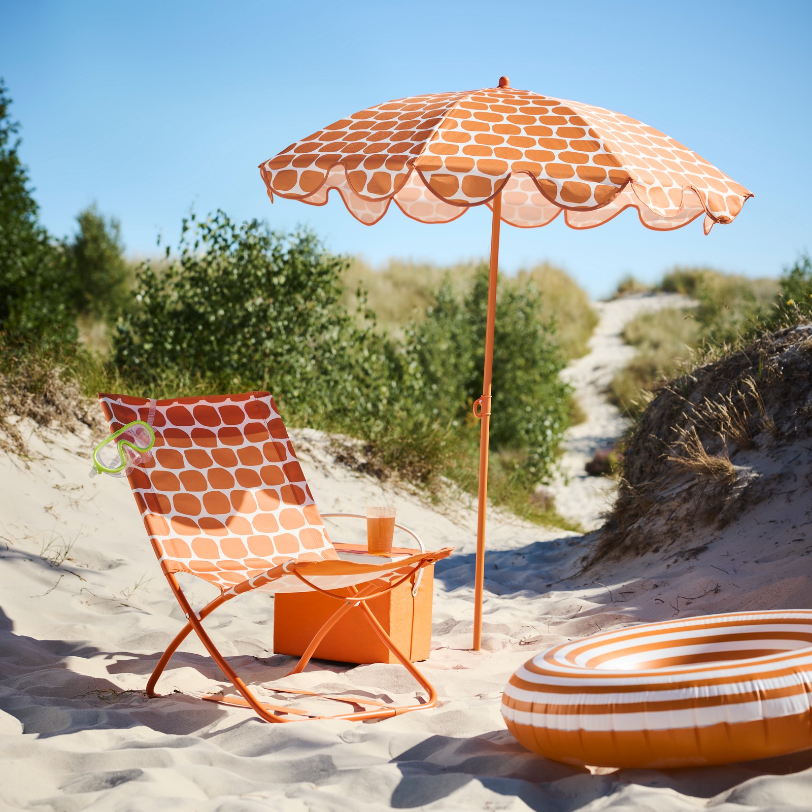 A beach scene with a bright orange STRANDÖN parasol and a beach chair with matching dotted pattern and an orange cooling bag.