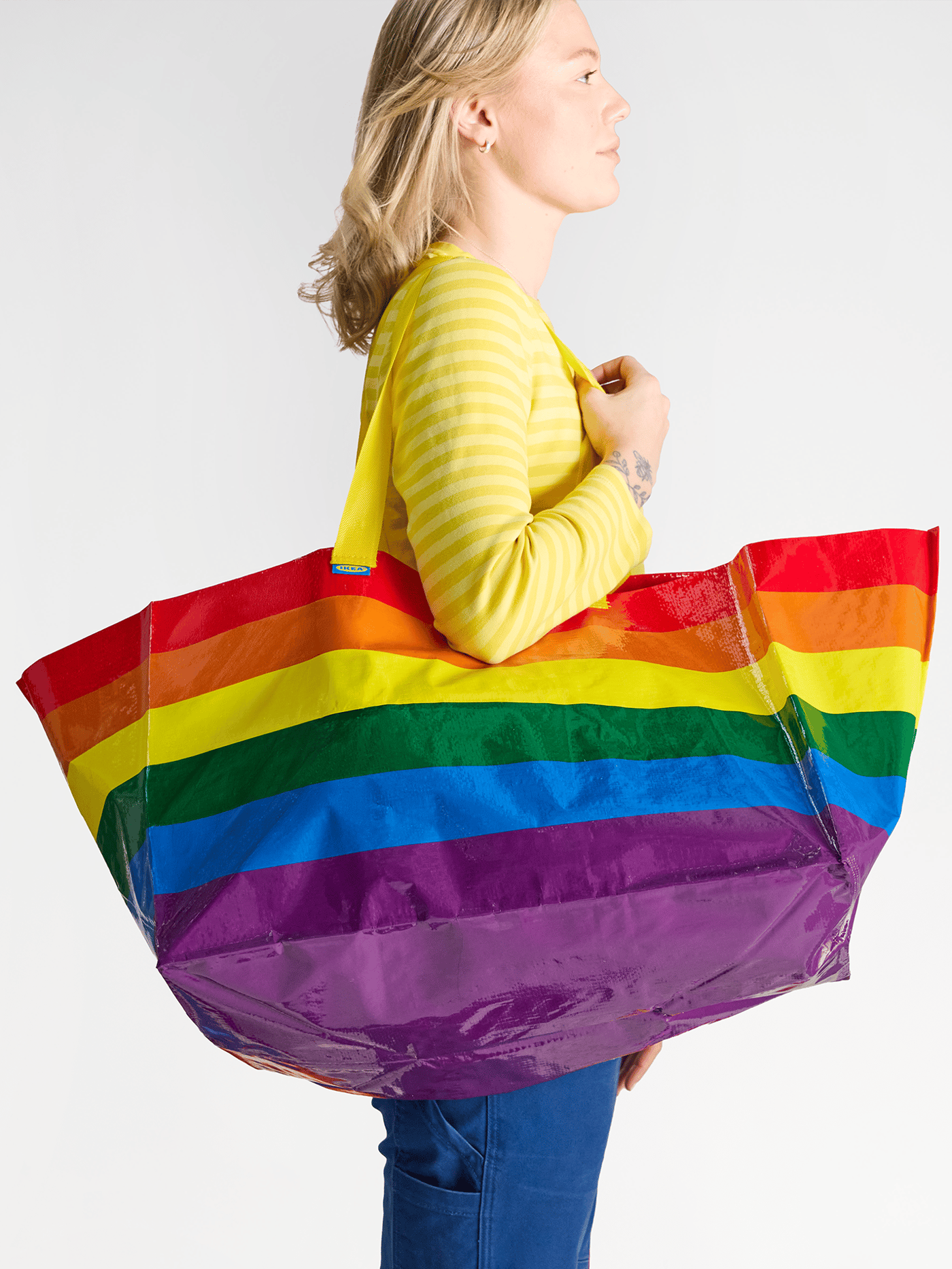 An IKEA co-worker wearing the IKEA uniform, holding a STORSTOMMA rainbow bag.