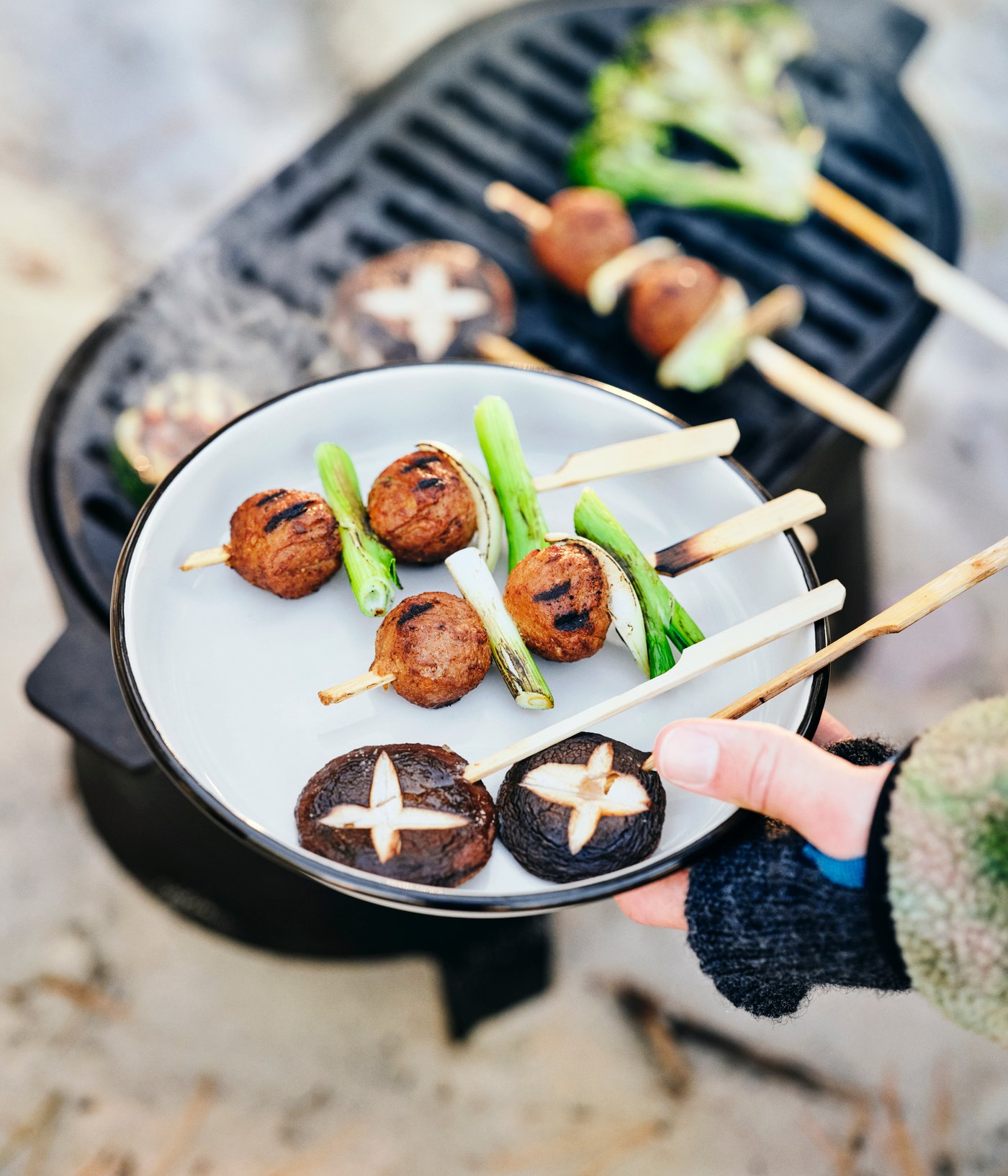 Una mano sostiene  un plato SOLUPPGÅNG con brochetas de setas y albóndigas vegetales,cerca    de una barbacoa SOLUPPGÅNG.