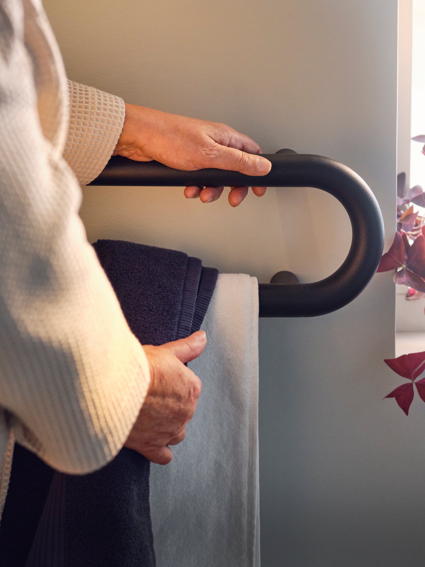 A person is holding onto a BÄSINGEN towel rail in anthracite with one hand and removing a grey towel with the other hand.