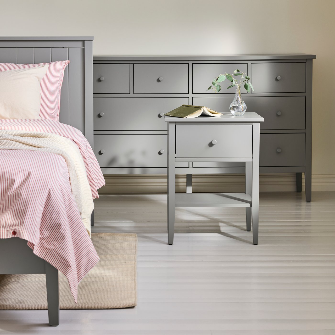 A GULLABERG bedside table in grey placed beside a bed with patterned bedding in a bedroom with soft, neutral tones.
