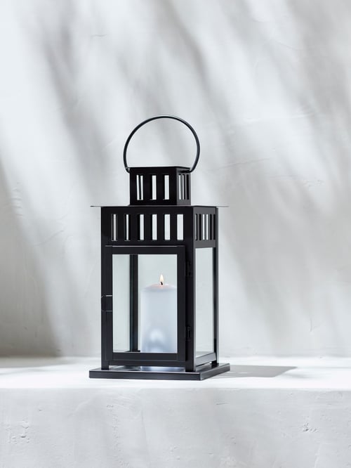A black, steel and glass BORRBY indoor/outdoor lantern holds a lit, white block candle. It is placed alone on a shaded step.
