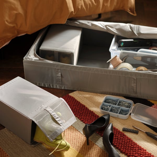 Dark grey SKUBB boxes/baskets are on a multicolor/red BRÖNDEN rug by a bed and filled with tools and shoes in a bedroom.