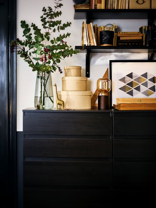Two KULLEN chests of five drawers with the top filled with decorative objects. Dark, wall-mounted shelves above.