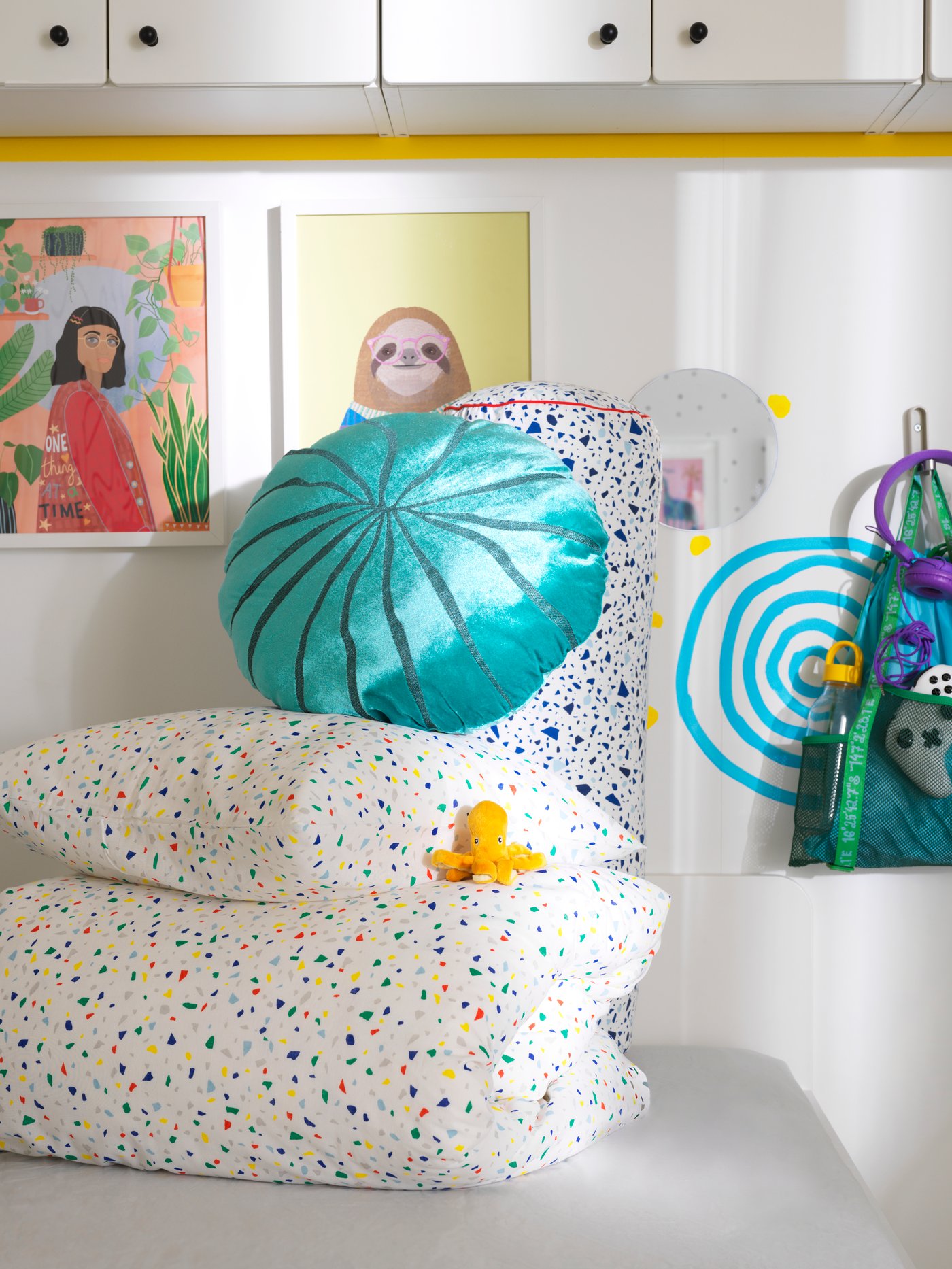 A coral-shaped, turquoise BLÅVINGAD cushion sits on top of a pillow and duvet in MÖJLIGHET bed linen piled up on a bed.