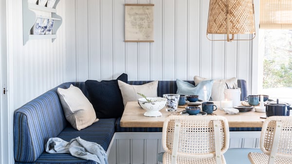 Ett ljust matrum med vit panelvägg och ett hörnplacerat sittbänkssystem med blå och beige textilier. På bordet står porslin i mörkblått och vitt, glas, en kanna, skålar och tallrikar, samt en mindre vas med blommor. Bordet är av ljust trä och framför det står två stolar med flätade rottingsitsar. Ovanför bordet hänger en lampskärm i naturmaterial. På väggen bakom soffan sitter en enkel hylla med porslin samt en inramad karta eller illustration. Ett fönster till höger släpper in naturligt ljus och visar ett ljust utomhuslandskap. Flera runda markörer är placerade över bilden som interaktiva punkter.