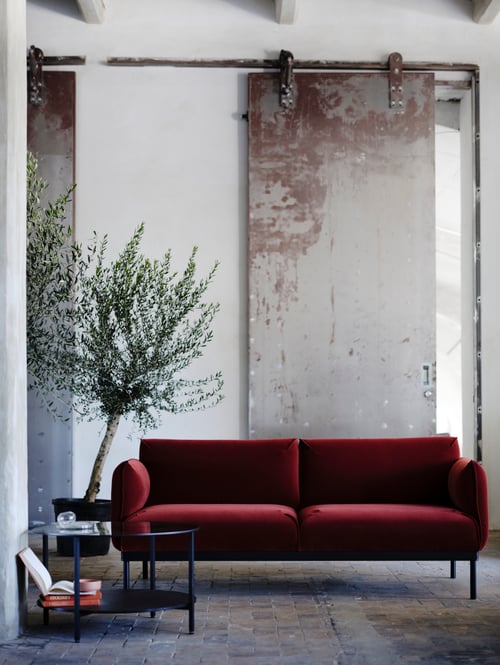 Une causeuse ÄPPLARYD rouge-brun dans un espace industriel et rustique avec une table basse et un arbre en arrière-plan.