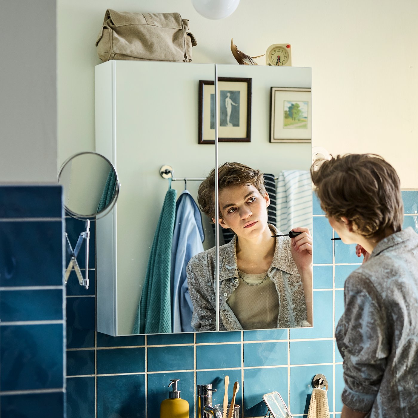 A person in a blue-tiled bathroom looks at their reflection in an ENHET mirror cabinet with two doors.