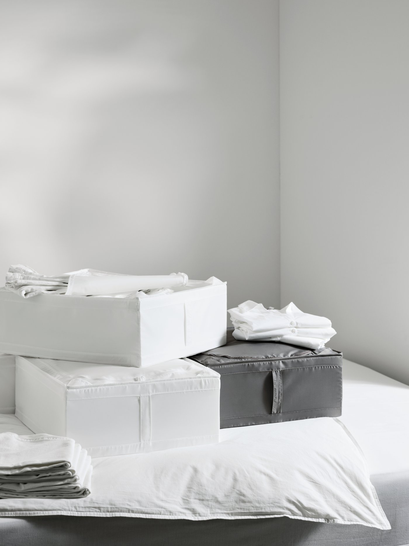 Dua kotak storan SKUBB berwarna putih dipenuhi dengan tekstil di atas satu sama lain, di sebelah kotak SKUBB berwarna kelabu. Semua berada di atas katil.
