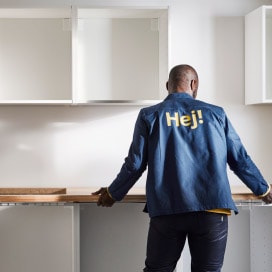 En person i blå jakke med teksten "HEJ" på ryggen står foran kjøkkenbenk og hvite veggseksjoner.