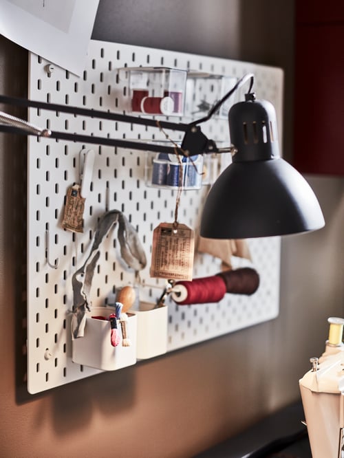 Arbeitsbereich mit weißem SKÅDIS Lochblech, verschiedenem Bastelbedarf und einer an der Wand montierten TERTIAL Arbeitsleuchte mit verstellbarem Arm.
