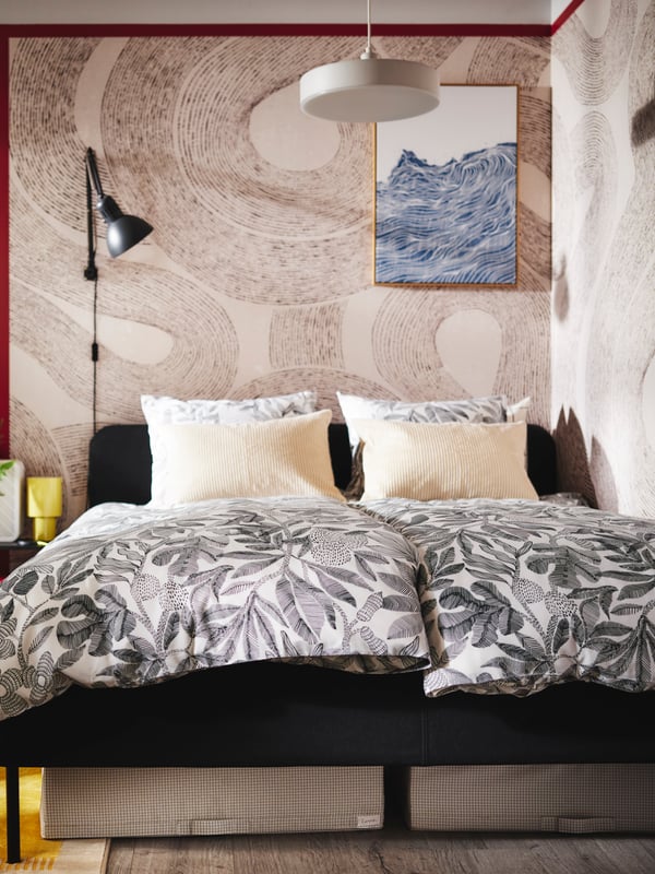 A dark grey SLATTUM upholstered bed topped with two duvets covered in BROKDRACENA bed linen stands in the corner of a bedroom.