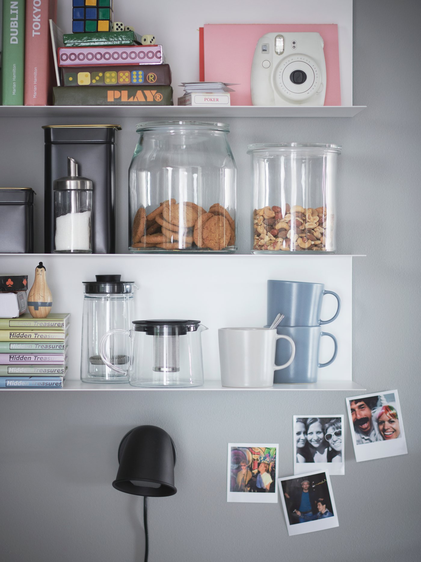 Vägghyllor med glasburkar fyllda med kakor och müsli, muggar, böcker, kamera och små foton. Nedanför sitter en svart vägglampa.
