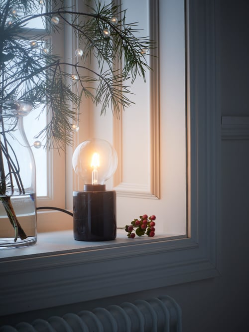 A LUNNOM LED clear bulb fitted in a MARKFROST marble table lamp, standing on a windowsill next to pine branches in a vase.