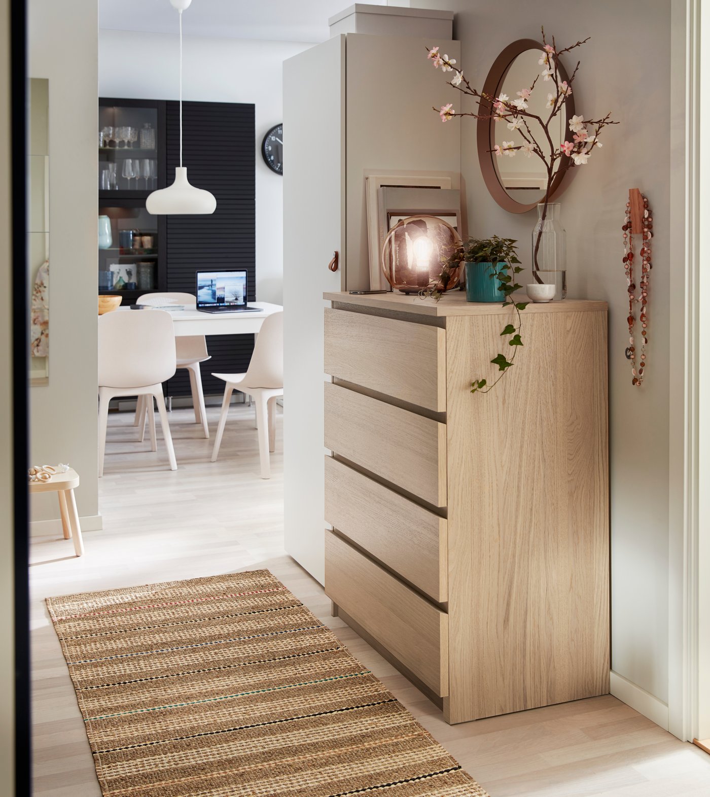 En MALM träbyrå med spegel, växt och dekorationer står i en korridor med vävd matta. I bakgrunden syns en matplats med stolar, ett bord med en laptop ovanpå, samt mörka skåp och hyllor längs väggen.