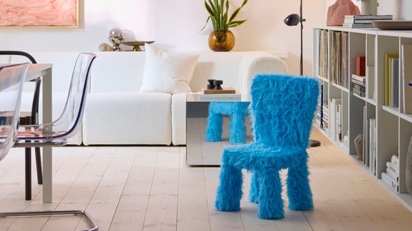 En barnstol i ett GRESIMOJS stolsöverdrag i blå fuskpäls står i ett ljust och modernt vardagsrum, bredvid ett sidobord med spegel.