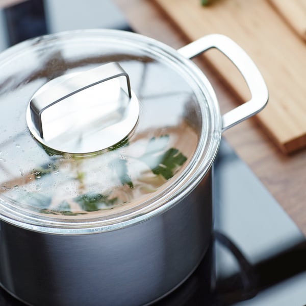 A stainless-steel pot with a glass lid on an induction cooktop next to a wooden cutting board.