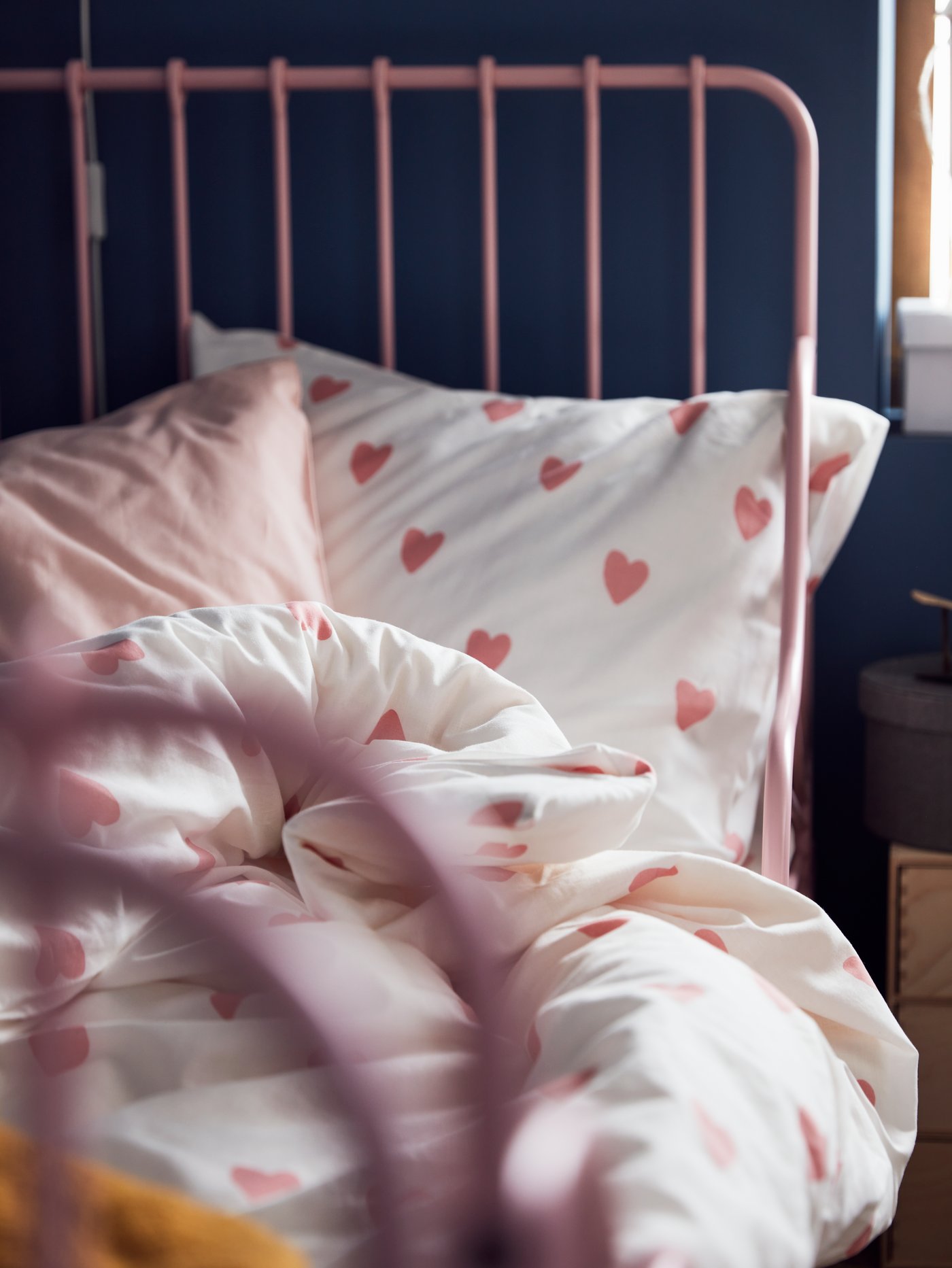 Cama rosa claro con ropa de cama con estampado de corazones en blanco/rosa, junto al borde de una minicómoda de contrachapado de abedul.
