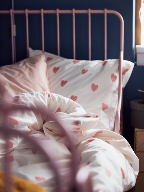 A light pink bed made with heart patterned white/pink bed linen, by the edge of a birch plywood mini chest of drawers.