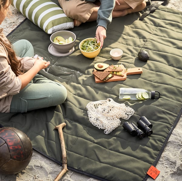 Deux personnes sont assises sur une couverture matelassée verte lors d'un pique-nique sur la plage, avec des collations et des boissons. Un oreiller rayé, un bâton en bois et un ballon de football se trouvent à proximité, créant une atmosphère détendue et chaleureuse.