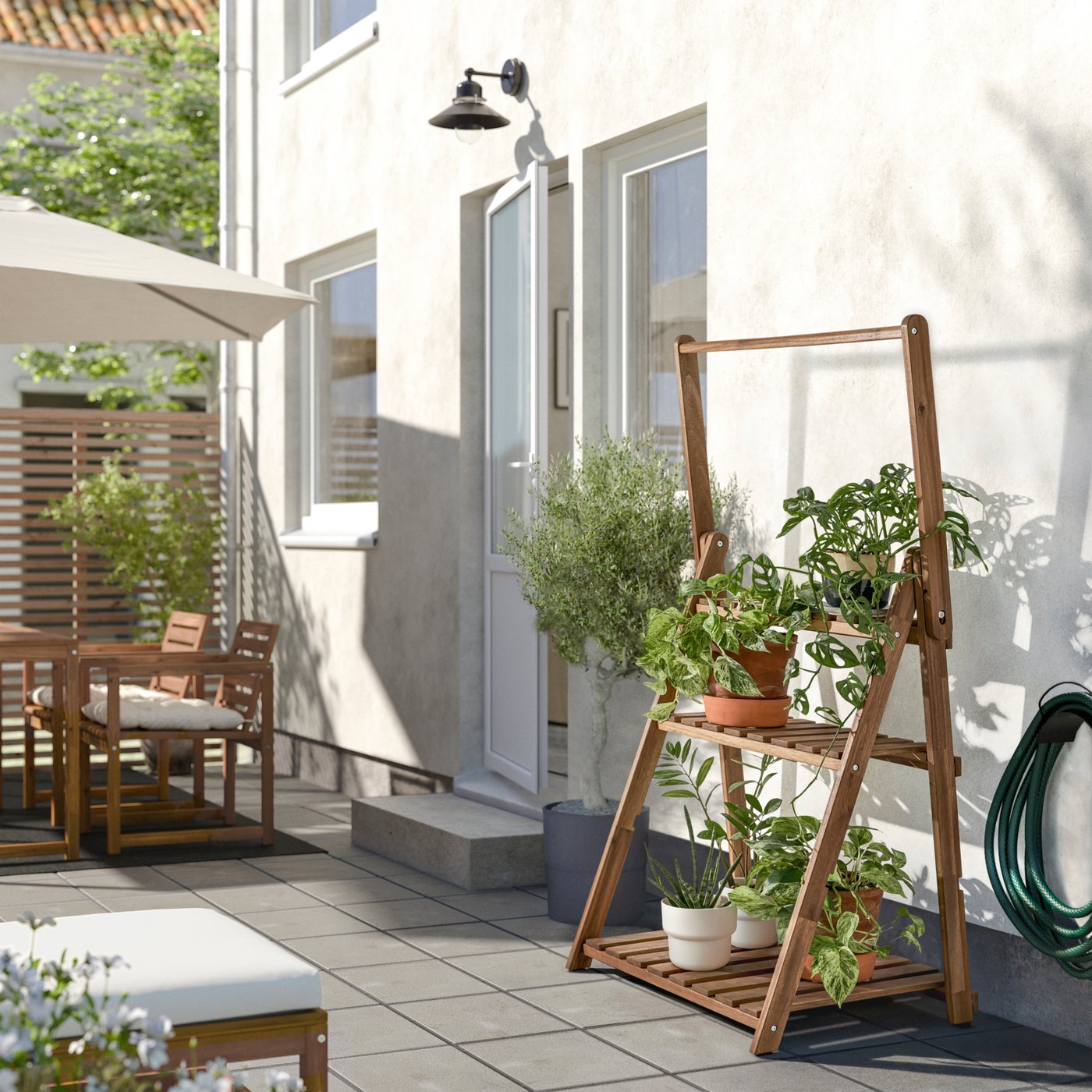 Utemiljö med matplats under parasoll, växter på träställning, glasdörr i vit fasad och trädgårdsslang på husväggen. 

