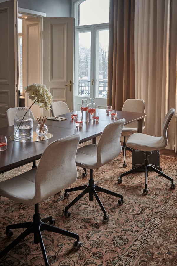 Elegant mötesrum med beige stolar runt ett träbord, dekorerat med blommor och glas. Stora fönster med gardiner och en mönstrad matta på golvet.