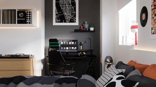 Bedroom with a bed in the foreground and a desk by a wall painted black in the back, with a computer and gaming chair.