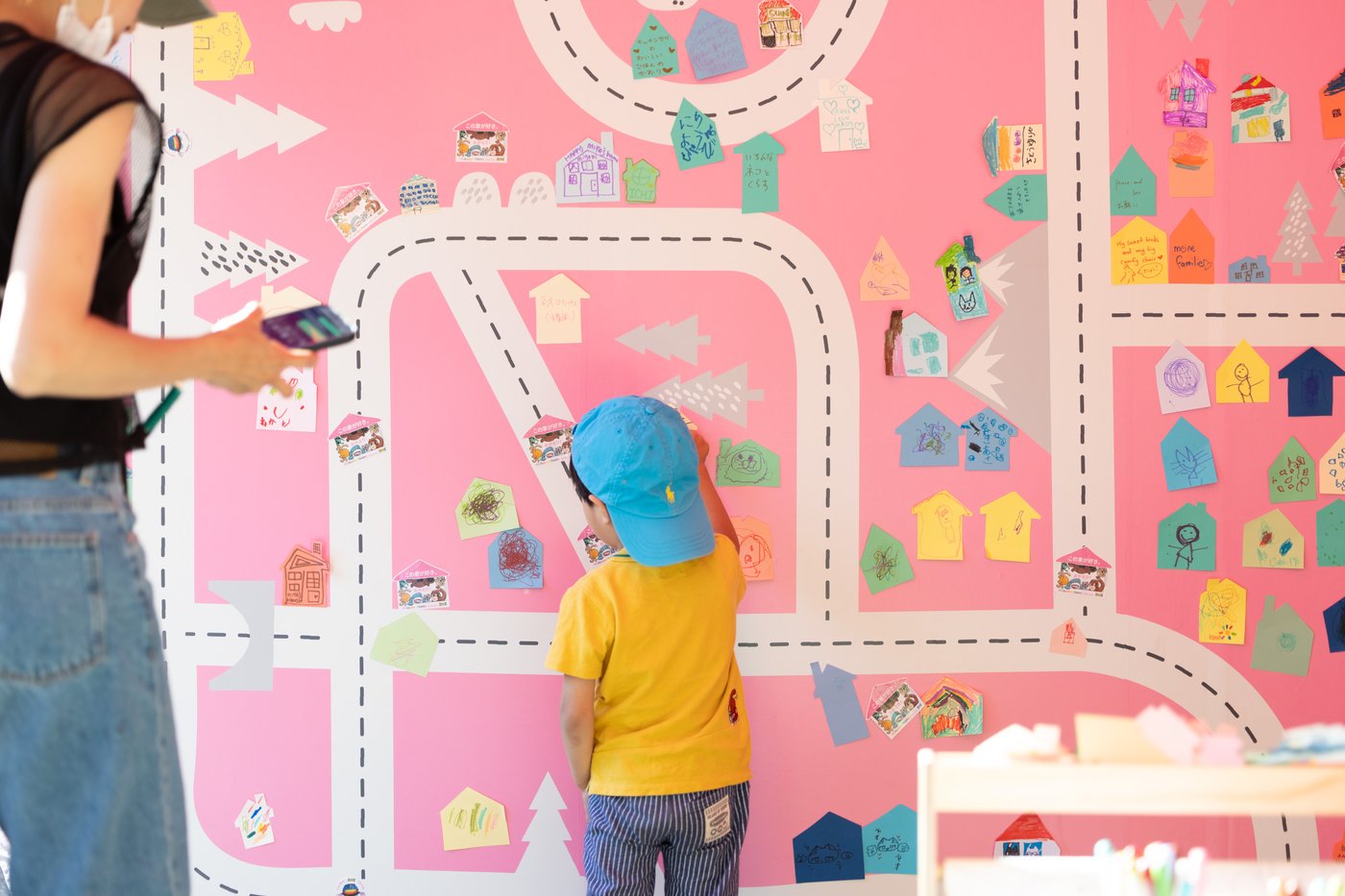 ピンクの壁に家やメッセージのカラフルなカードが並び、子どもがイケアのWelcome Homeイベントで自分のカードを貼っている。
