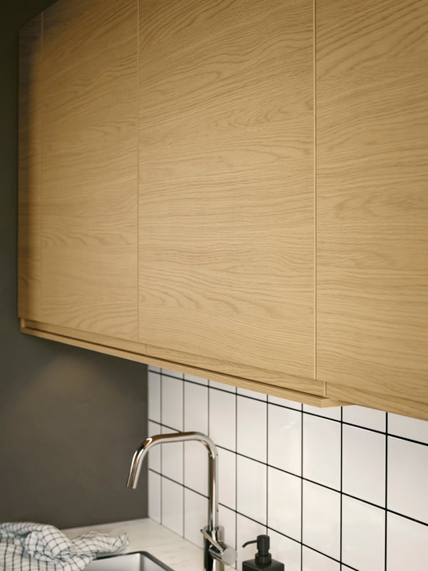 Pensili con frontali VOXTORP effetto rovere con maniglie integrate posizionati al di sopra di un lavello con un miscelatore.