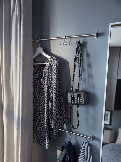 Schlafzimmer mit zwei KUNGSFORS Stangen an der Wand. Daneben sind zwei HANNALILL Gardinenschals und ein HOVET Spiegel zu sehen. An den Stangen hängen Accessoires und Kleidung auf einem Bügel.