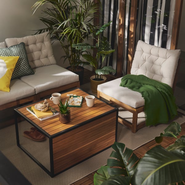 A lush balcony with a versatile light brown HÅKANSKÄR coffee table and seating surrounded by potted plants and soft cushions.