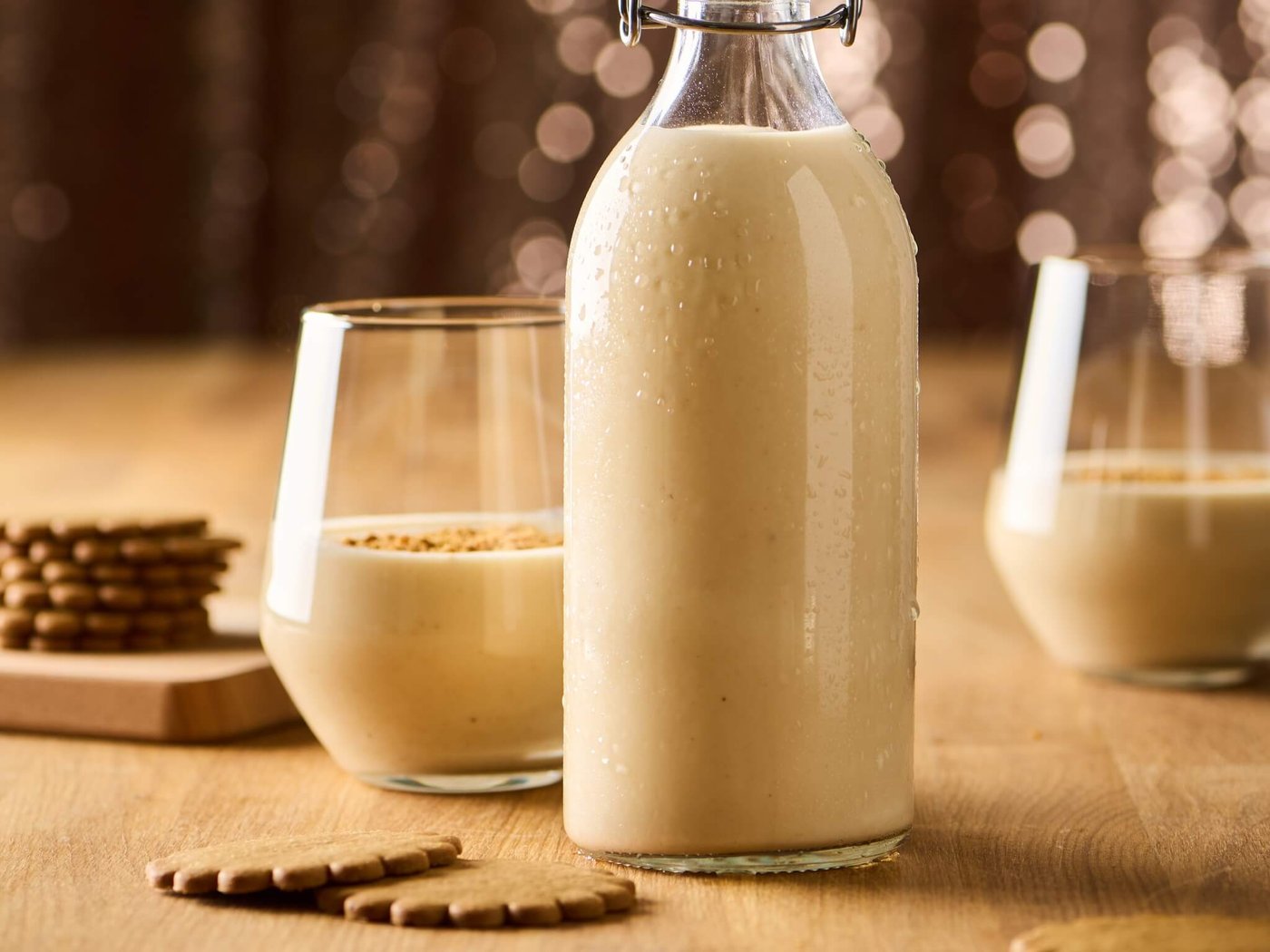 Creamy coquito in a glass bottle on a wooden table, with two filled glasses and cookies arranged around it.