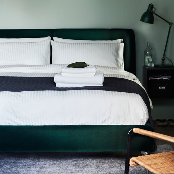 A green bedroom with a dark green upholstered bed covered in white bed linen. A work lamp is on a bedside table.