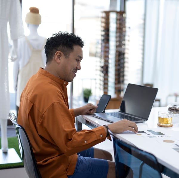 Eine Person sitzt vor einem Laptop an einem Tisch und hält ein Smartphone in der Hand.