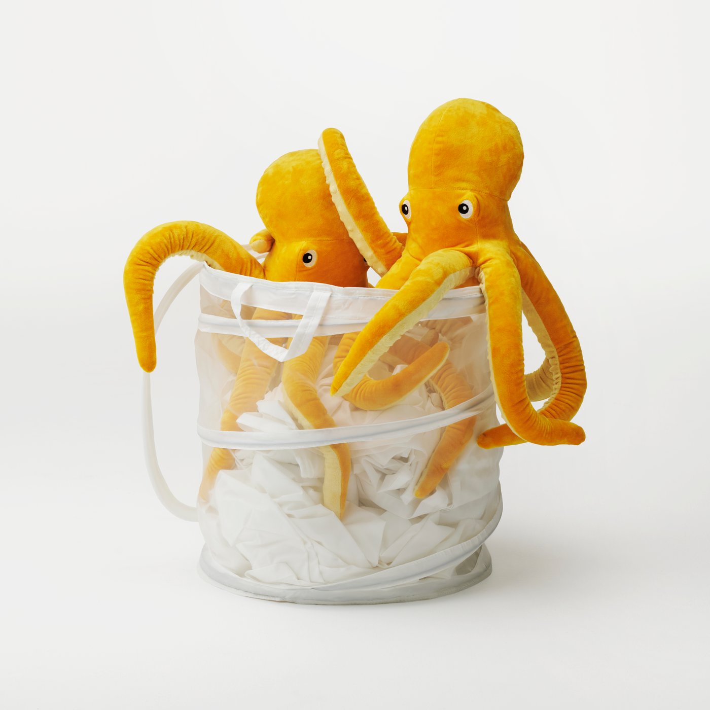 Two yellow BLÅVINGAD octopus soft toys sitting on top of white laundry in a basket in a white studio setting.
