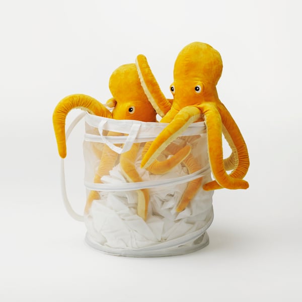 שני צעצועי תמנון צהובים של BLÅVINGAD מונחים על גבי כביסה לבנה בסל בסטודיו לבן.