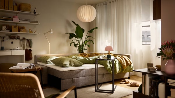 Es ist ein FRIHETEN Bettsofa im Bettmodus in einem sanft beleuchteten Wohnzimmer mit Gardinen und Rollos zu sehen, die größtenteils geschlossen sind.