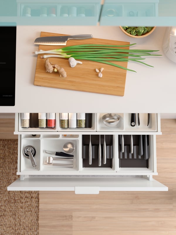 An anthracite-coloured UPPDATERA cutlery caddy, cutlery and TREBENT chopsticks, next to a glass carafe and a stack of plates.