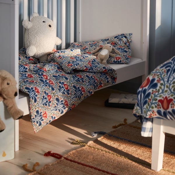 A white SUNDVIK cot with one long side removed has a floral SÄTERVIDE duvet cover and pillowcase and two soft toys inside.