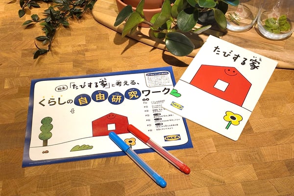 イケアのテーブルに、絵本「たびする家」と赤い家のイラストが描かれた自由研究ワークシート、赤と青のペン、グリーンの植物が並んでいる。