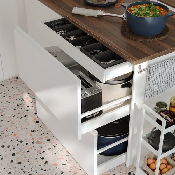 Several drawers in a kitchen peninsula with white VOXTORP fronts have been pulled out, revealing small appliances inside.