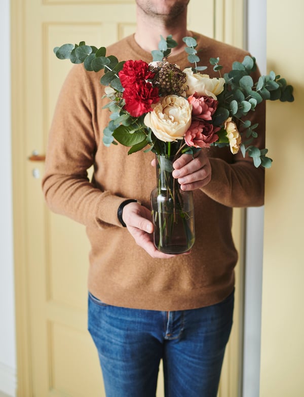 Eine Person in Jeans und einem Pullover, die eine FÖRENLIG Vase mit SMYCKA Kunstblumen in den Händen hält.