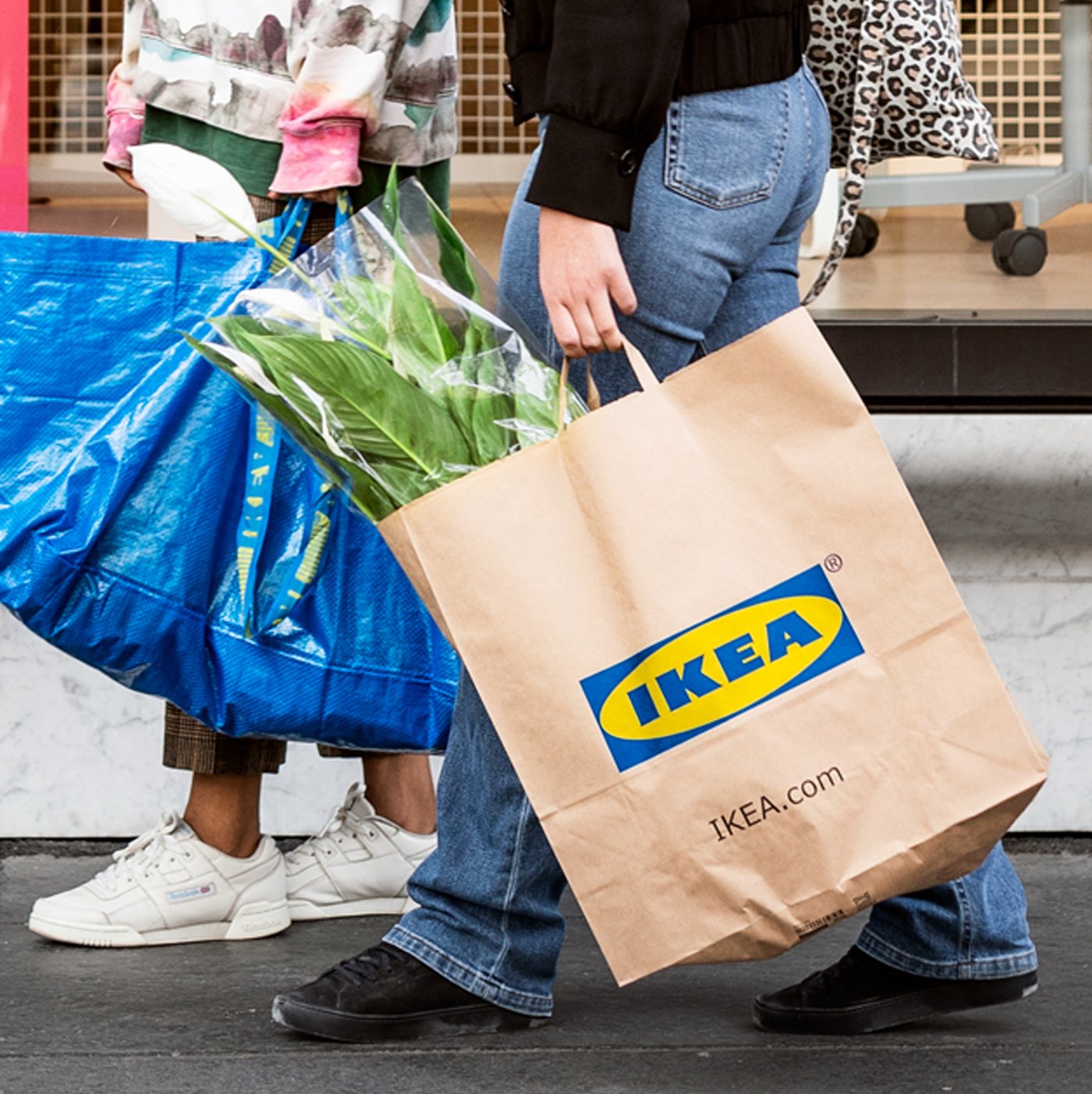 Two people who have been shopping at IKEA, one carrying a blue FRAKTA bag and the other an IKEA paper bag containing plants.