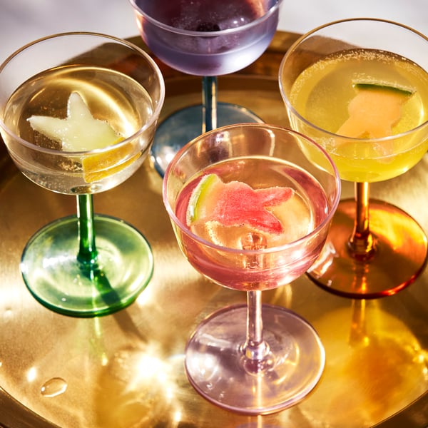 A set of multicolour TJÄRLEK glasses sit on a tray filled with drinks and fresh fruit garnish. 
