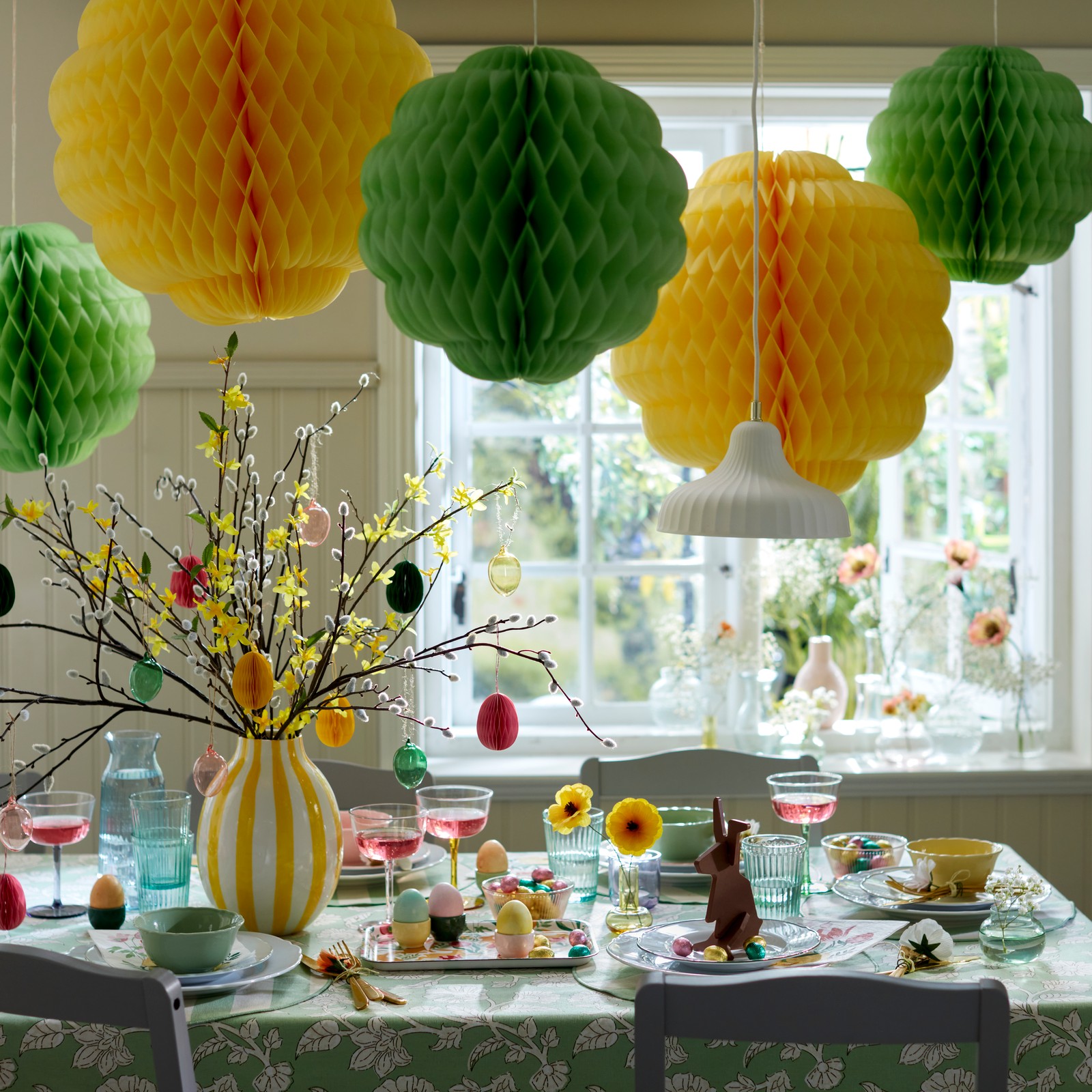 A bright dining table setting featuring mixed coloured TJÄRLEK glassware, surrounded by festive TJÄRLEK hanging decorations.