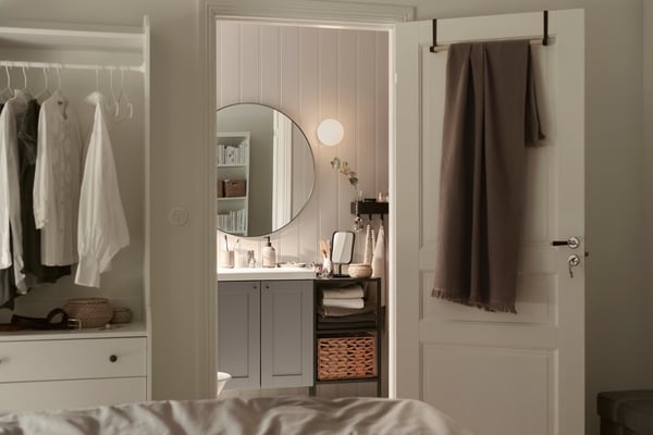 An ENHET/TVÄLLEN wash-basin cabinet with two doors with a round mirror above, in a small bathroom adjoining a bedroom.