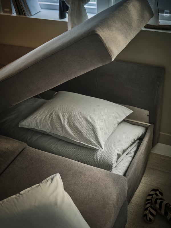 Close-up of extra quilt cover and pillows piled up inside the grey HOLMSUND sofa-bed. Part of the sofa is open for storage.